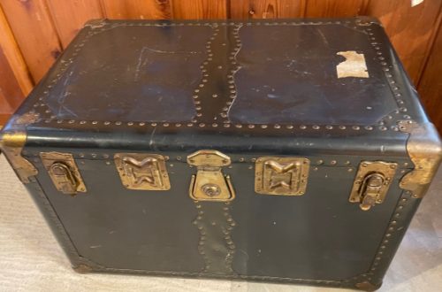 View of the top and front of the travel trunk. Black with gold clasps.